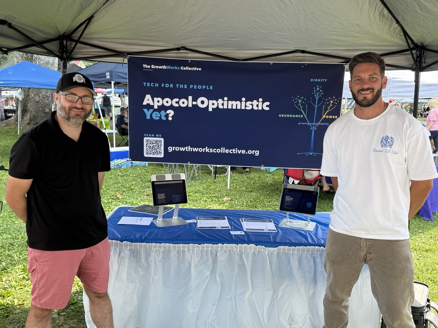 Two people at The GrowthWorks Collective booth at an outdoor community event, beside a table of tablets under a banner that reads Tech for the People.